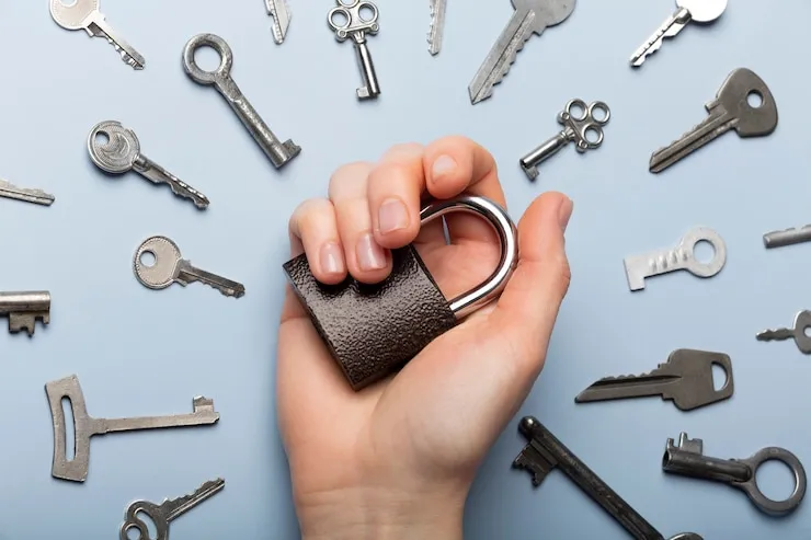 A hand holding an open padlock surrounded by an assortment of house and cabinet keys